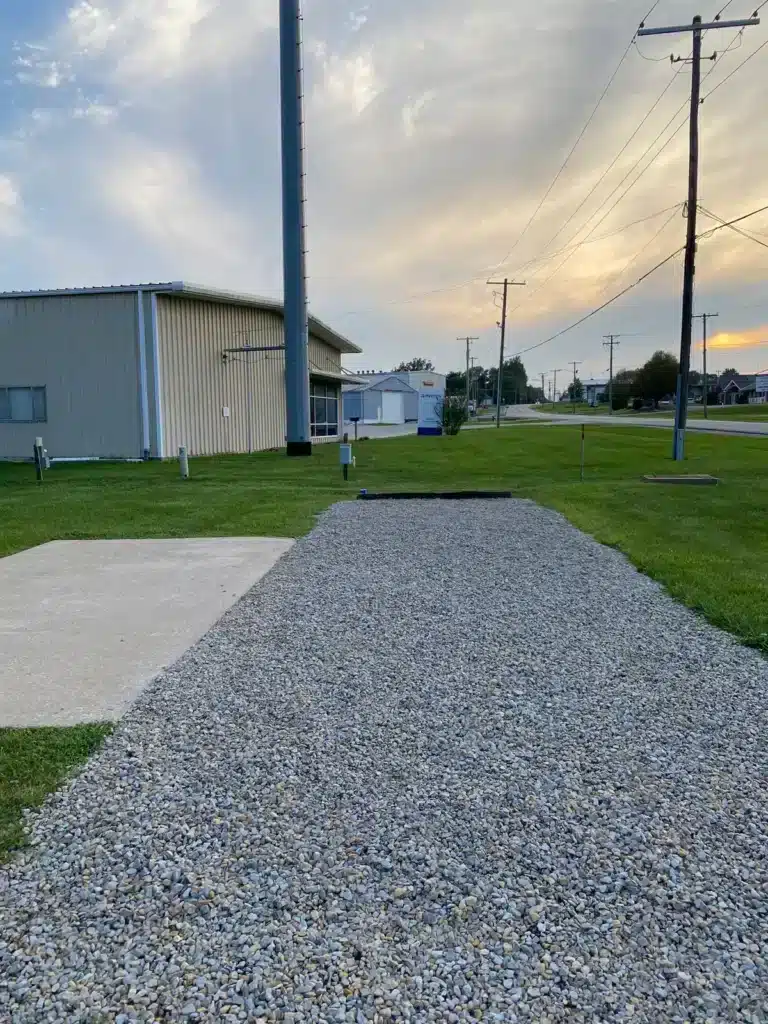 Empty gravel RV pad beside a concrete patio and grassy lawn with utility hookup pedestals, metal building, and sunset