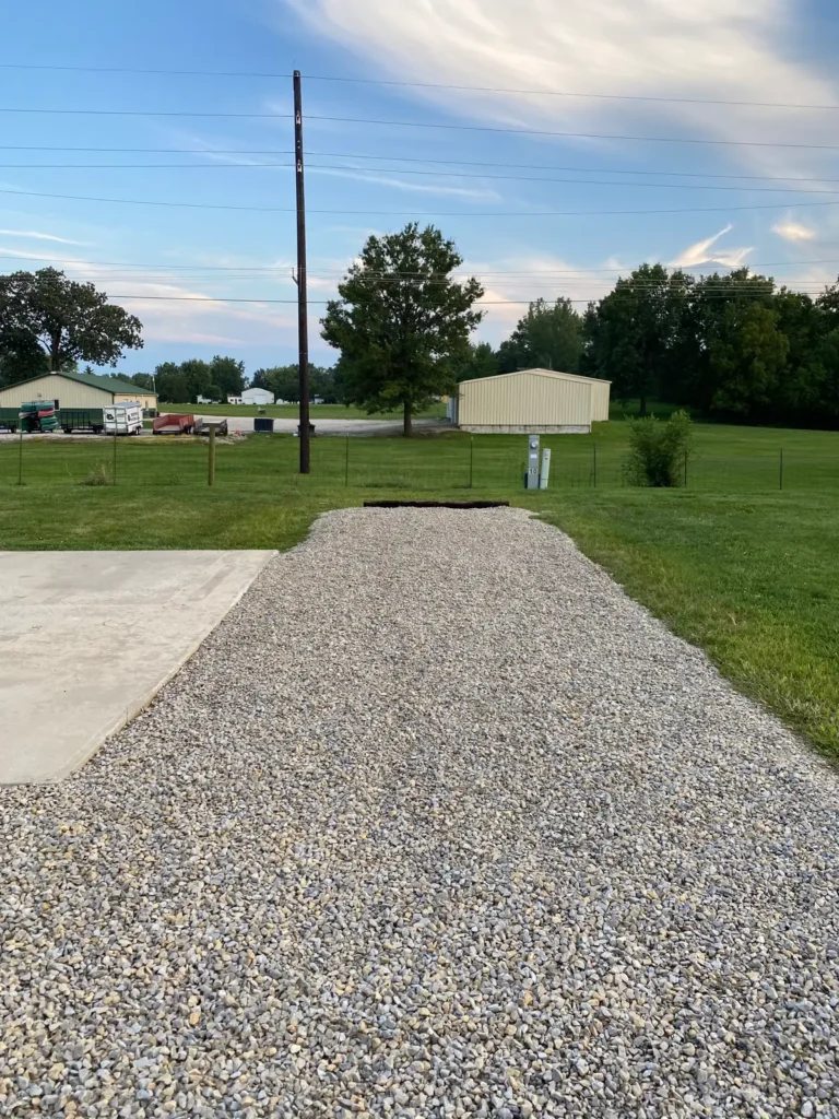 Empty gravel RV pad with adjacent concrete patio slab, utility hookup pedestal marked site 10, grass and metal barns