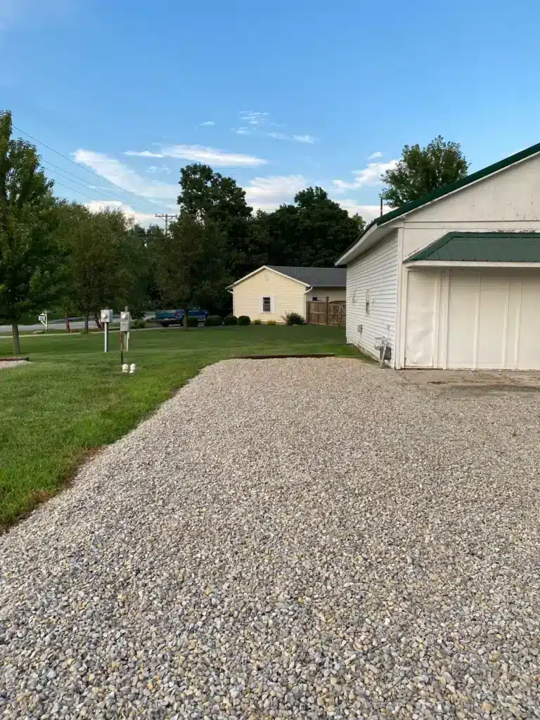 Wide gravel driveway pad beside a white metal garage with green roof, mowed lawn, utility pedestals and trees nearby.