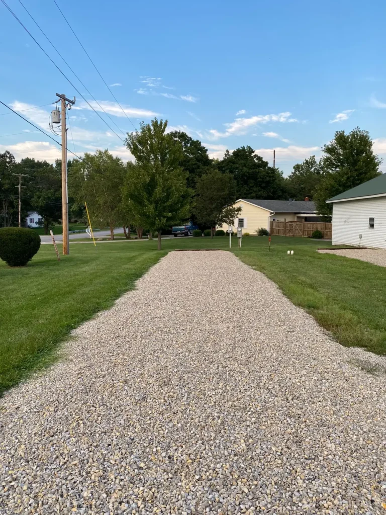 Wide gravel driveway leading toward a residential street, flanked by mown grass, utility pole, mailbox posts, and a