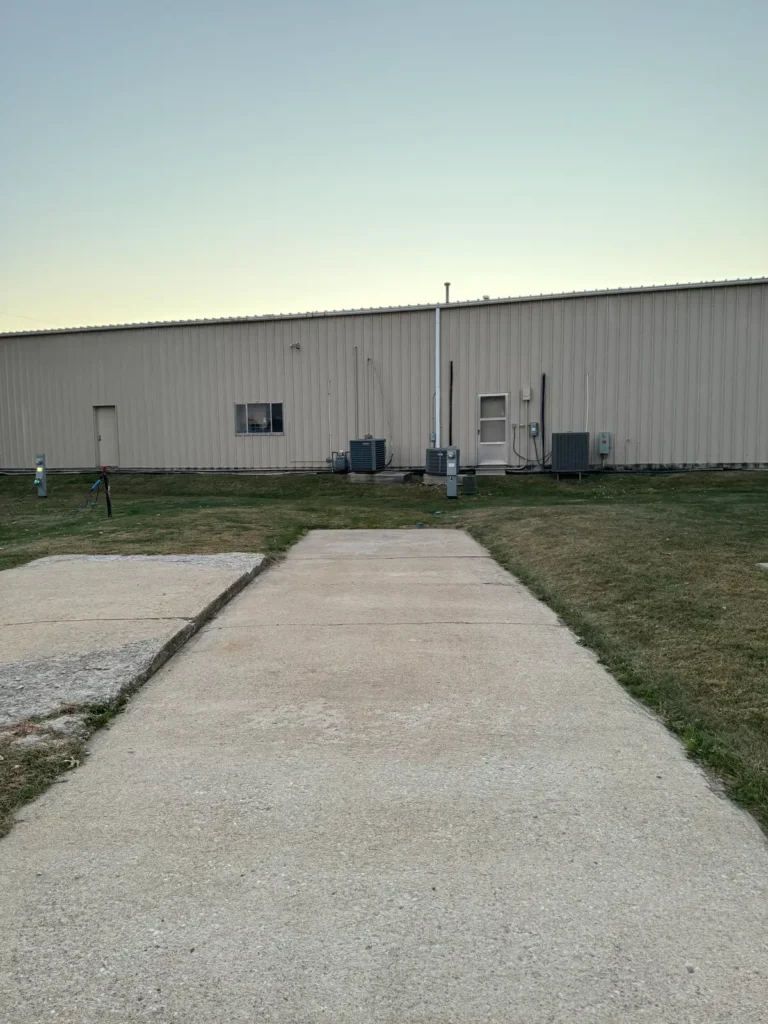 Wide concrete walkway leading to a tan metal building with a door, window, air conditioning units and utility pedestals.