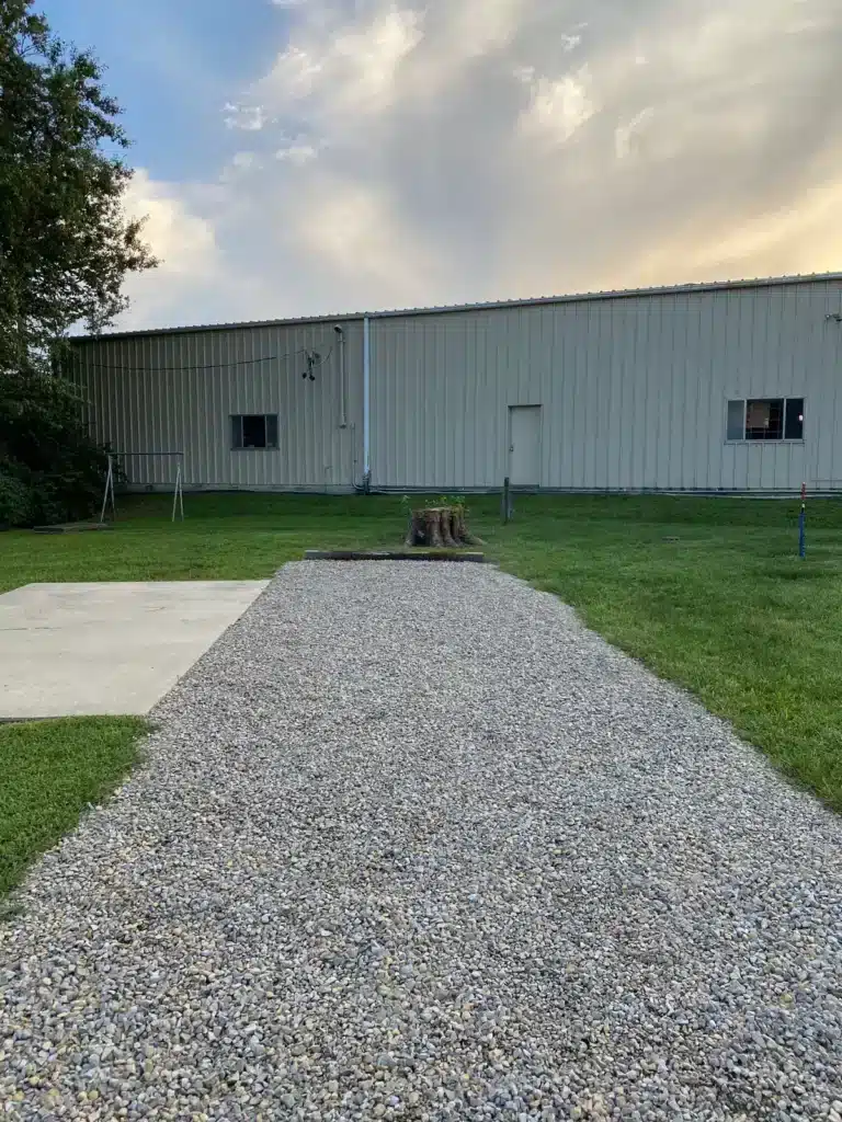 Gravel RV pad with adjacent concrete patio slab and tree stump, fronting a large tan metal building at dusk.