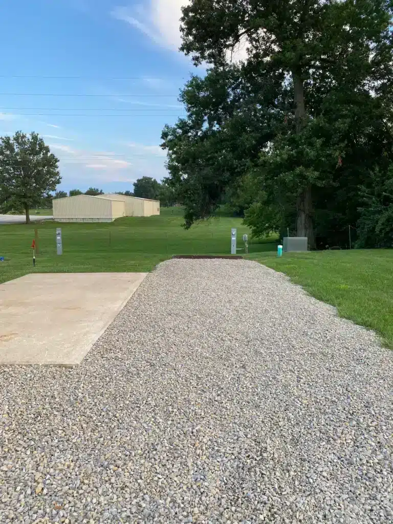 Empty gravel RV pad with adjacent concrete patio, numbered hookup pedestals, mowed lawn, large oak tree and metal