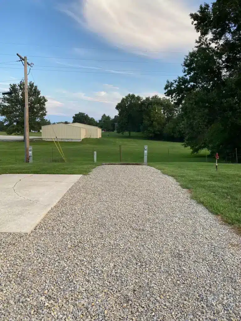 Empty gravel RV pad with adjacent concrete patio, numbered utility hookup pedestals, grassy field, power pole and metal