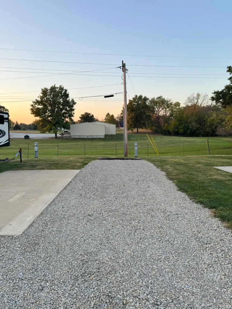 Empty gravel RV site with concrete patio, hookup pedestals, and utility pole, with trees and sunset sky beyond.