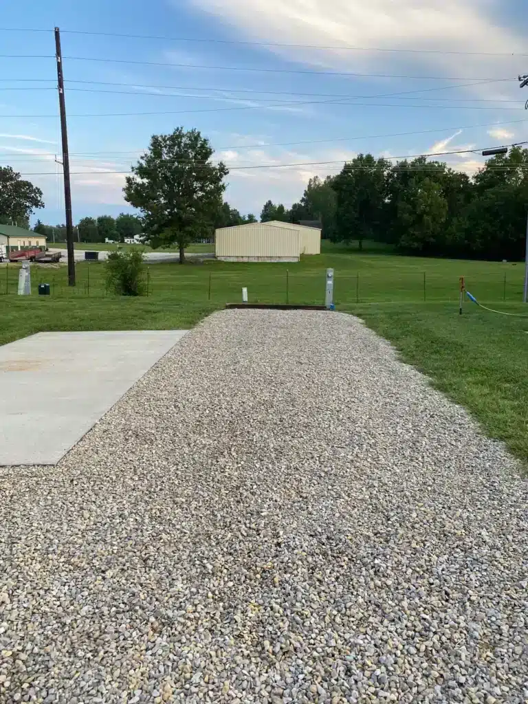 Empty gravel RV site with adjacent concrete patio pad, numbered utility pedestals with electric hookups, and a metal